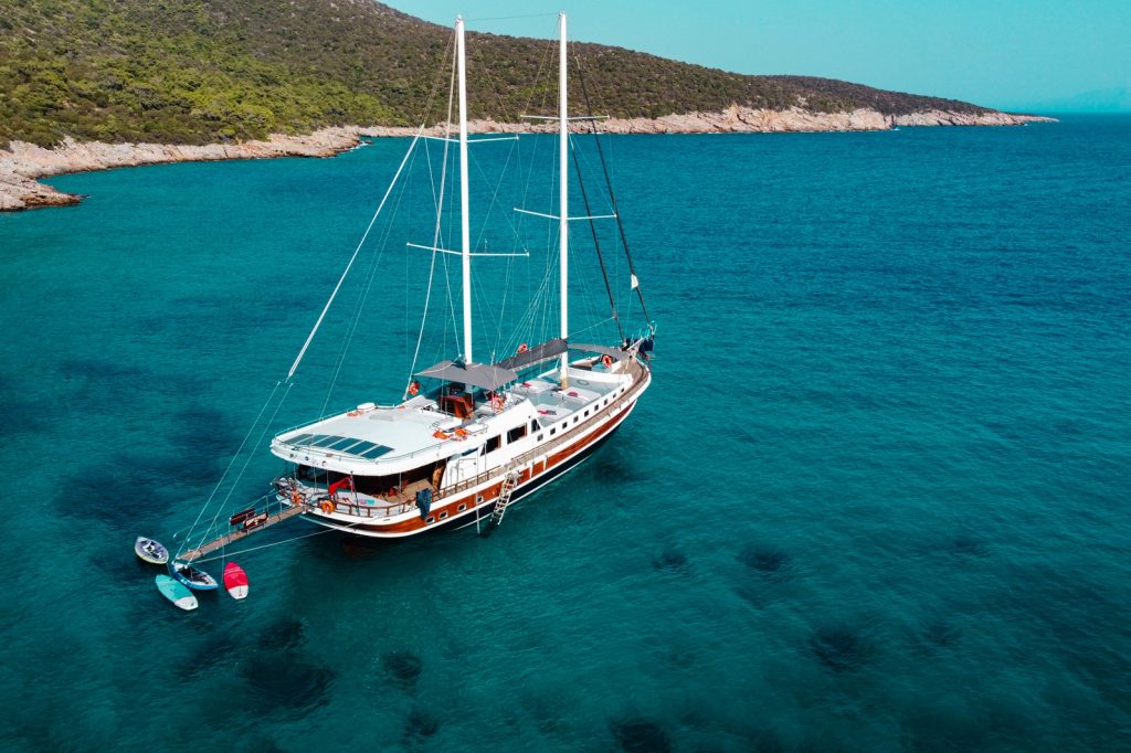 A large white and red yacht with two masts floats on clear blue water near a rocky, tree-covered shoreline under a clear sky. Paddleboards hang off the back of the yacht.