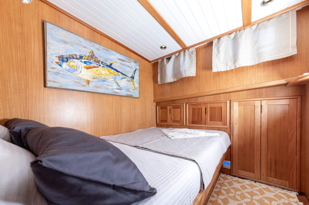 A cozy bedroom with wood-paneled walls and ceiling, a bed with white and dark gray bedding, a folded towel, and a large fish painting above the bed. Light enters through curtained windows on the right.