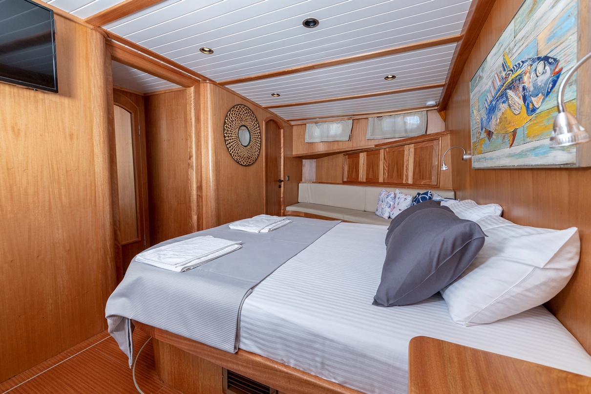 A cozy boat cabin with wooden walls, a large bed with gray and white linens, decorative pillows, a wall-mounted TV, a painting of a fish, and windows letting in natural light.