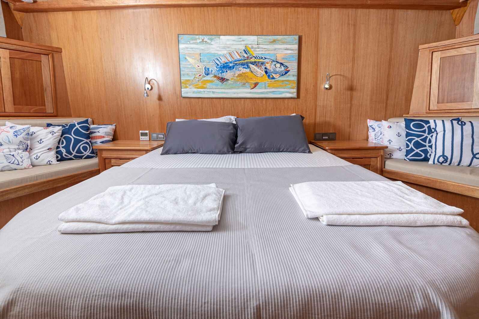 A neatly made bed with gray pillows and folded white towels, set in a cozy wooden room with nautical-themed cushions and a colorful fish painting on the wall above.