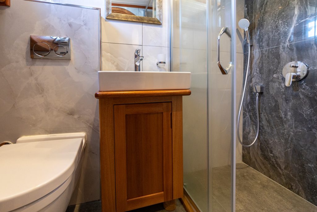 A small modern bathroom with a white toilet, a wooden vanity with a sink, a wall-mounted faucet, and a glass-enclosed shower with a handheld showerhead and gray marble wall tiles.