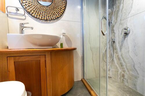 Modern bathroom with a wooden vanity, white vessel sink, round decorative mirror, glass shower enclosure, and marble-patterned shower wall. A liquid soap dispenser sits next to the sink.