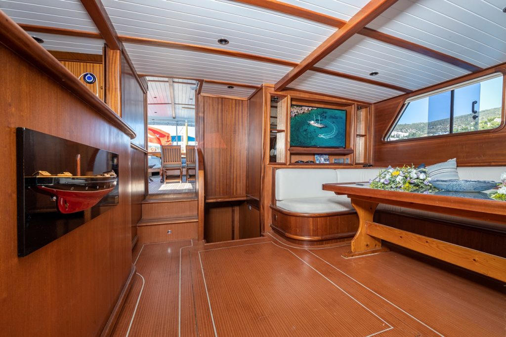 The interior of a wooden boat with polished floors, built-in bench seating around a dining table, a wall display with a boat model, and windows showing water and hills outside.