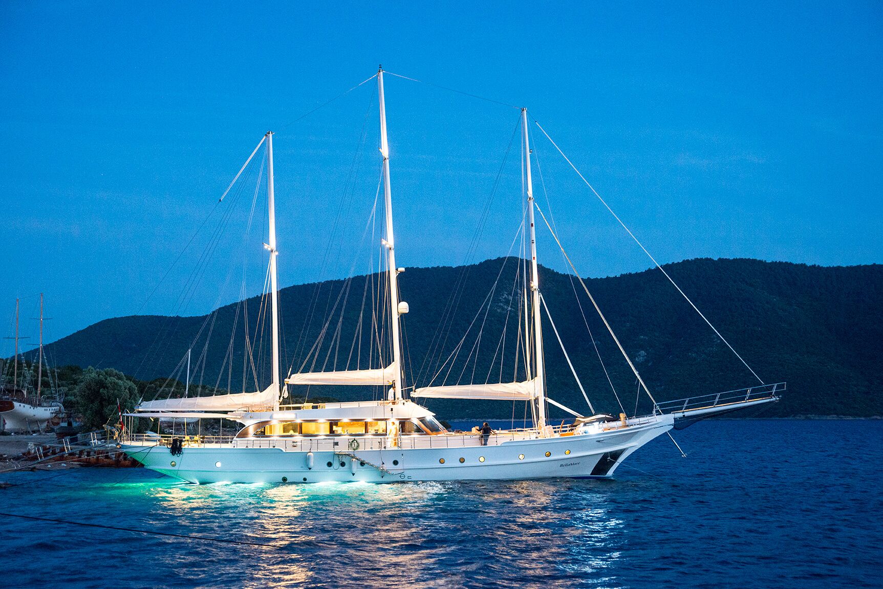 A large white yacht with three masts is illuminated and docked near a coastline at dusk, with dark hills and a deep blue sky in the background.