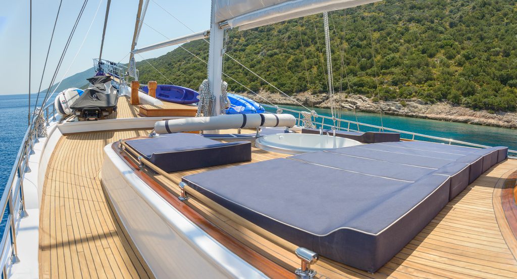 The deck of a luxury yacht with cushioned sunbeds, a hot tub, and water equipment, overlooking clear turquoise water and a green, tree-covered hillside.