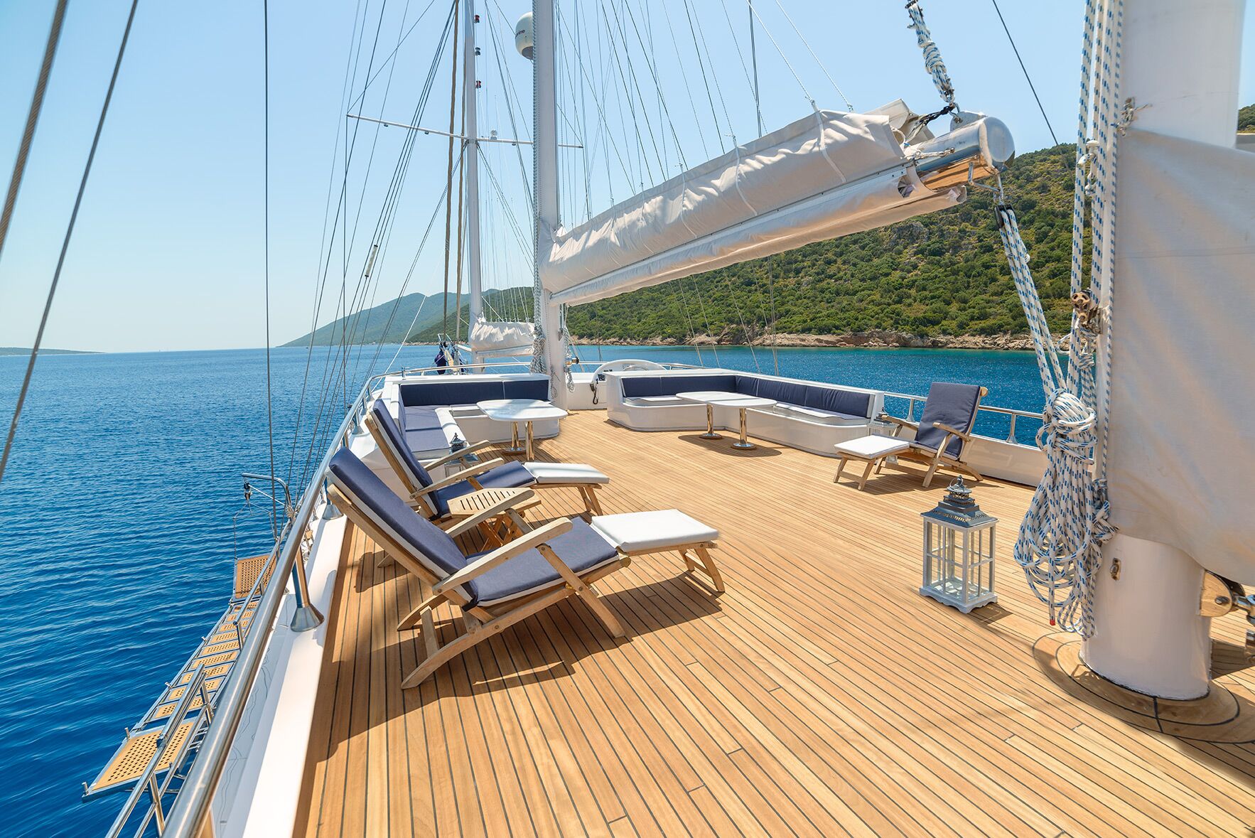 Sunny yacht deck with lounge chairs, tables, and cushioned seating overlooking calm blue sea and green hills on the coast; a lantern and rigging lines are visible under a clear sky.