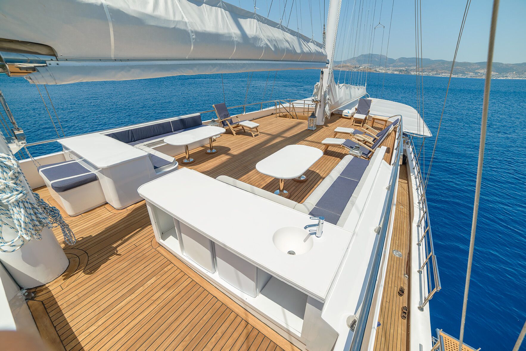 Spacious yacht deck with lounge chairs, tables, built-in benches, and a sink, overlooking calm blue sea and distant coastline under a clear sky.