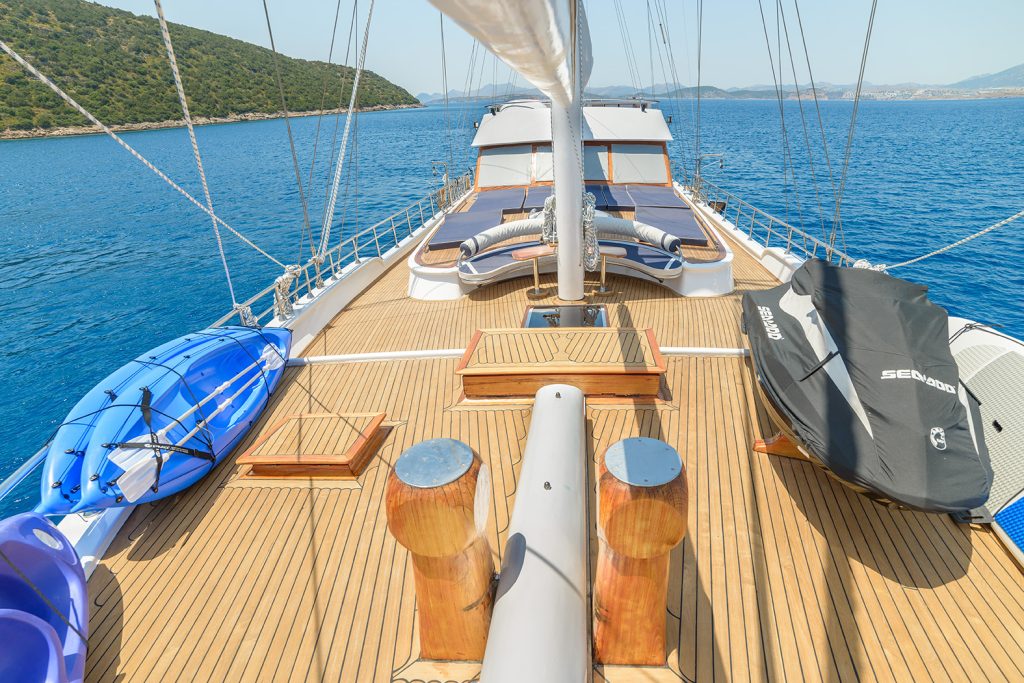 View from a yacht’s deck with lounge chairs, a kayak on the left, and a jet ski on the right. The yacht is sailing on calm blue water near a green, hilly coastline under a clear sky.