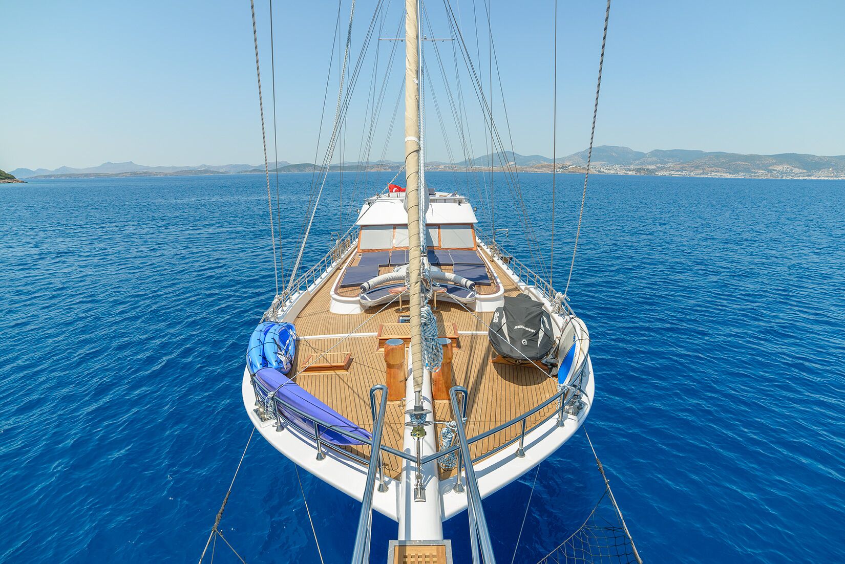 View from the bow of a sailing yacht on clear blue water, with sun loungers and seating on deck, surrounded by calm sea and distant hills under a clear sky.