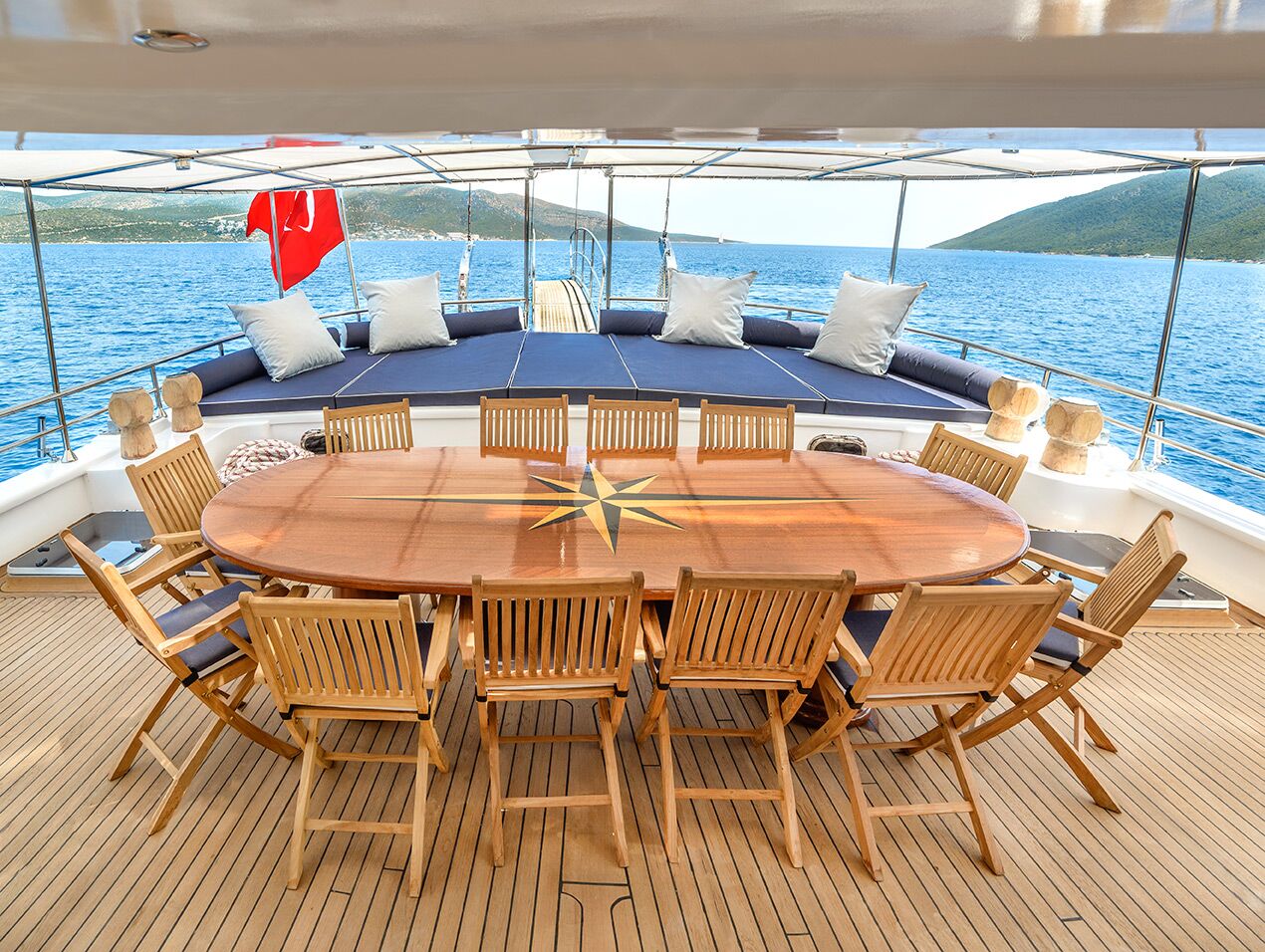 A large wooden dining table with twelve chairs is set on the deck of a yacht, overlooking the sea. Cushioned lounge chairs and a red flag are visible at the back, with distant green hills in the background.