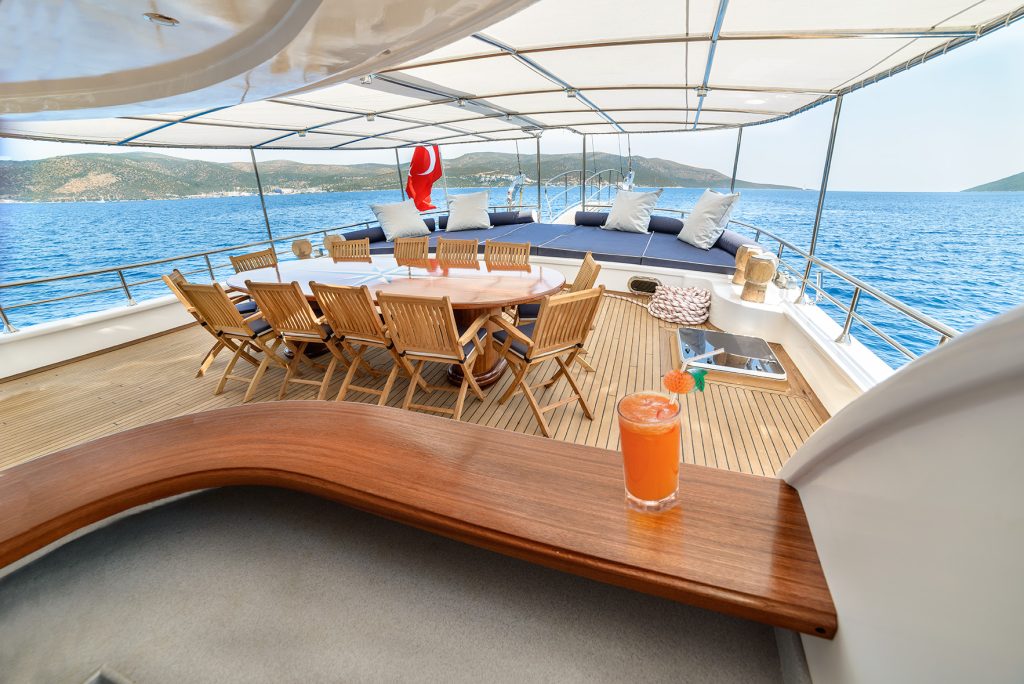 A luxury yacht deck with a wooden dining table and chairs, cushioned lounge area, and a glass of orange cocktail on a counter. The yacht is sailing on calm blue sea with hills in the background.