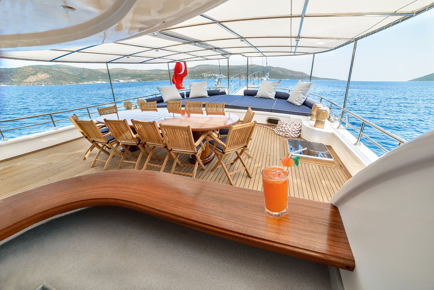 A luxury yacht deck with a wooden dining table and chairs, cushioned lounge area, and a glass of orange cocktail on a counter. The yacht is sailing on calm blue sea with hills in the background.