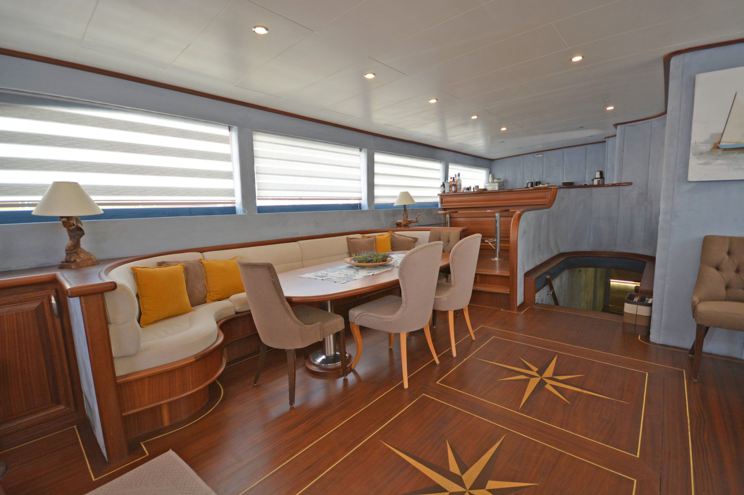 A yacht interior with wood flooring, a curved cushioned bench with yellow pillows, a round dining table, four beige chairs, and a bar area in the background. Large windows with striped blinds line the wall.