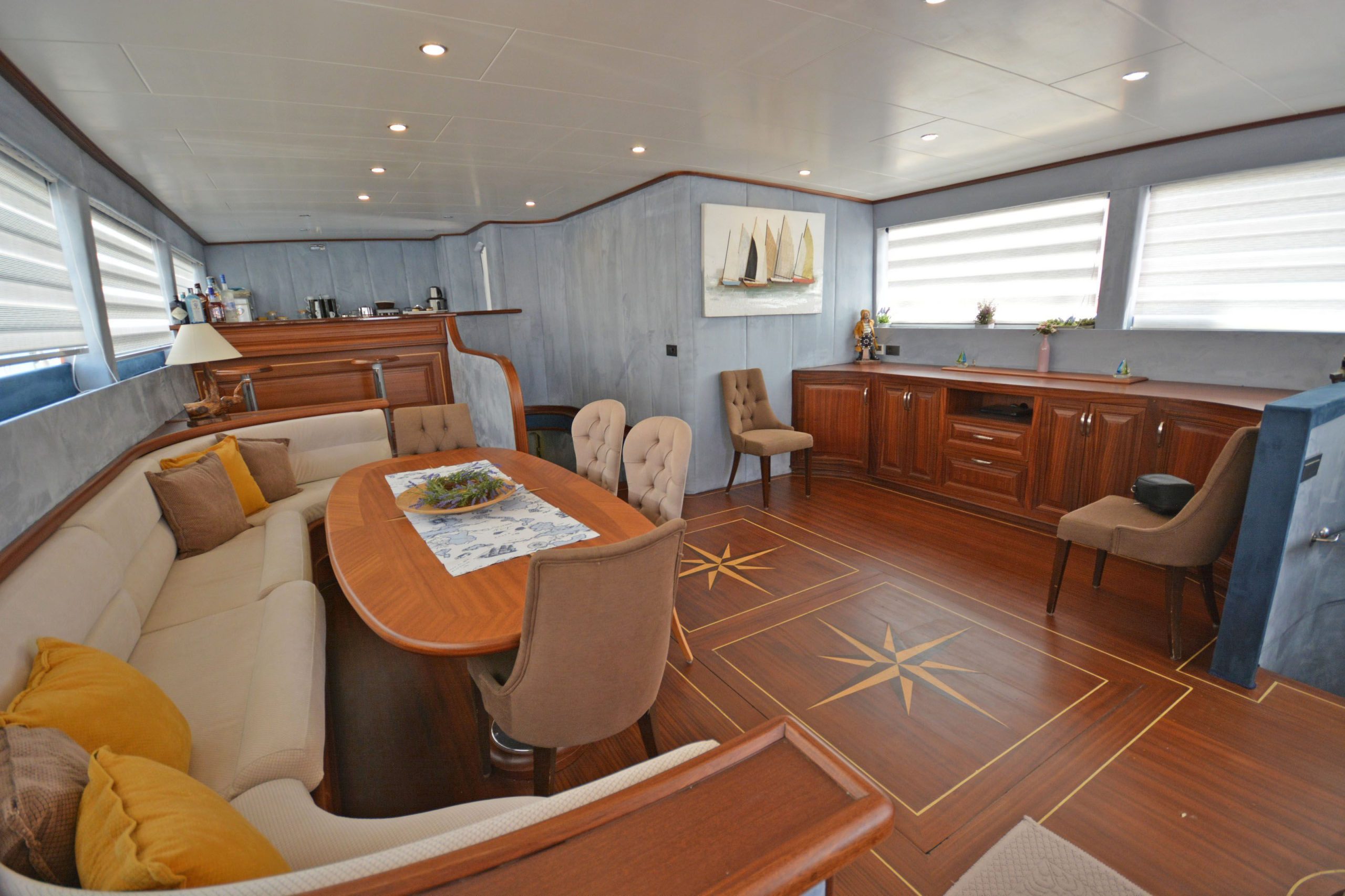 A yacht interior with wood flooring, a dining table with cushioned chairs, a beige sofa with yellow pillows, a cabinet, and nautical decor including a painting of sailboats and compass rose designs on the floor.