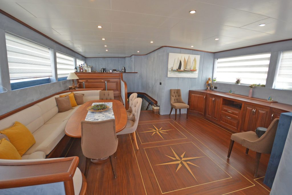 Spacious yacht interior with wood flooring, a dining area with beige chairs and cushions, built-in cabinetry, nautical decor, large windows with blinds, and a painting of sailboats on the wall.