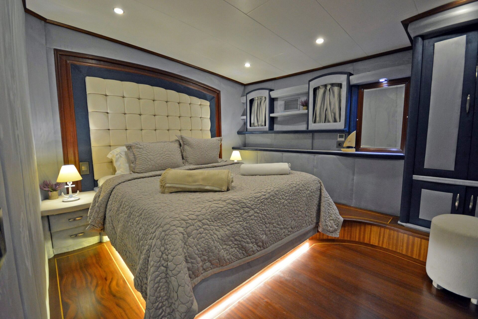 Luxurious bedroom with a large, neatly made bed, quilted gray bedding, padded headboard, wooden floor with under-bed lighting, built-in shelves, cabinets, and soft ambient ceiling lights.