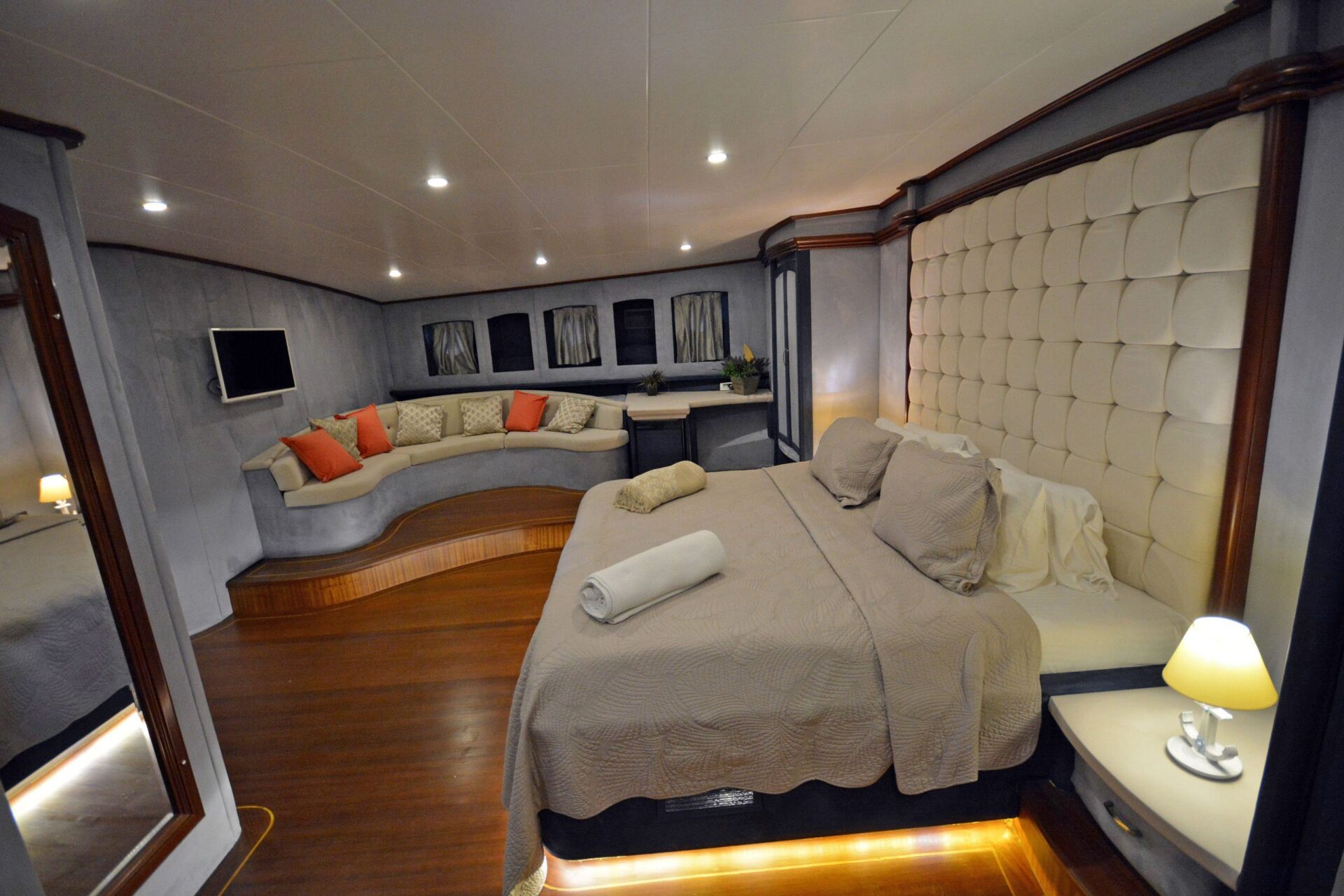 A spacious, modern bedroom with a large bed, beige bedding, and a padded headboard. There is a cozy sofa with orange pillows, wood flooring, a wall-mounted TV, and soft lighting throughout the room.