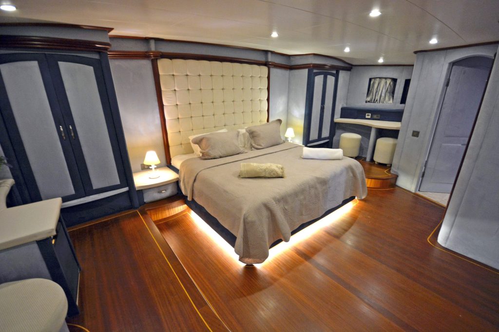 A spacious, modern bedroom with wood flooring, a large bed with beige linens, built-in wardrobes, bedside tables with lamps, and soft lighting under the bed and ceiling.