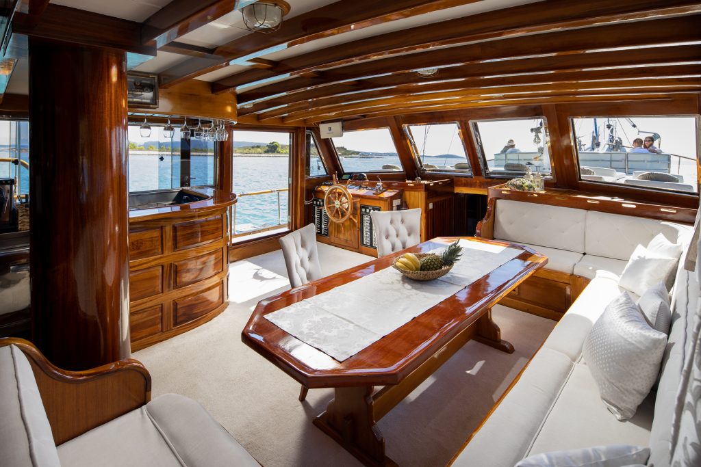 Luxury yacht interior with polished wood, white upholstered seating, a long dining table with a fruit bowl, large windows offering water views, and sunlight filling the elegant, spacious room.