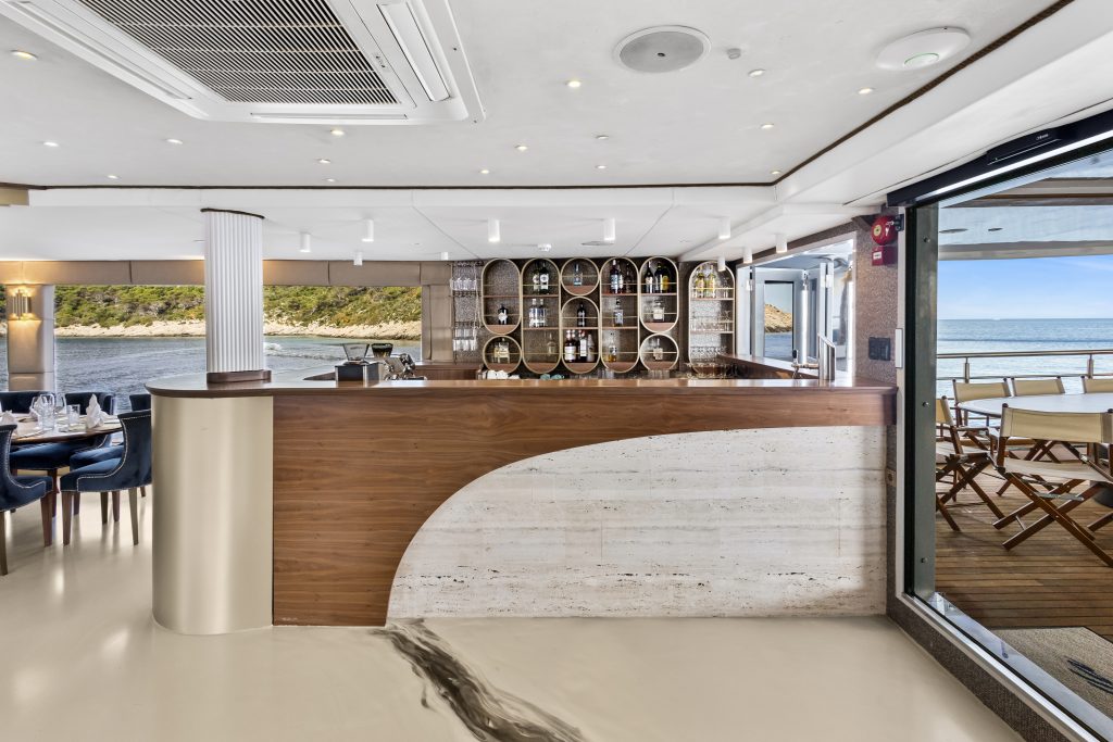 A modern indoor bar with wood and marble accents, shelves displaying bottles, and large windows opening to an outdoor terrace with tables and chairs overlooking the sea and a green coastline.