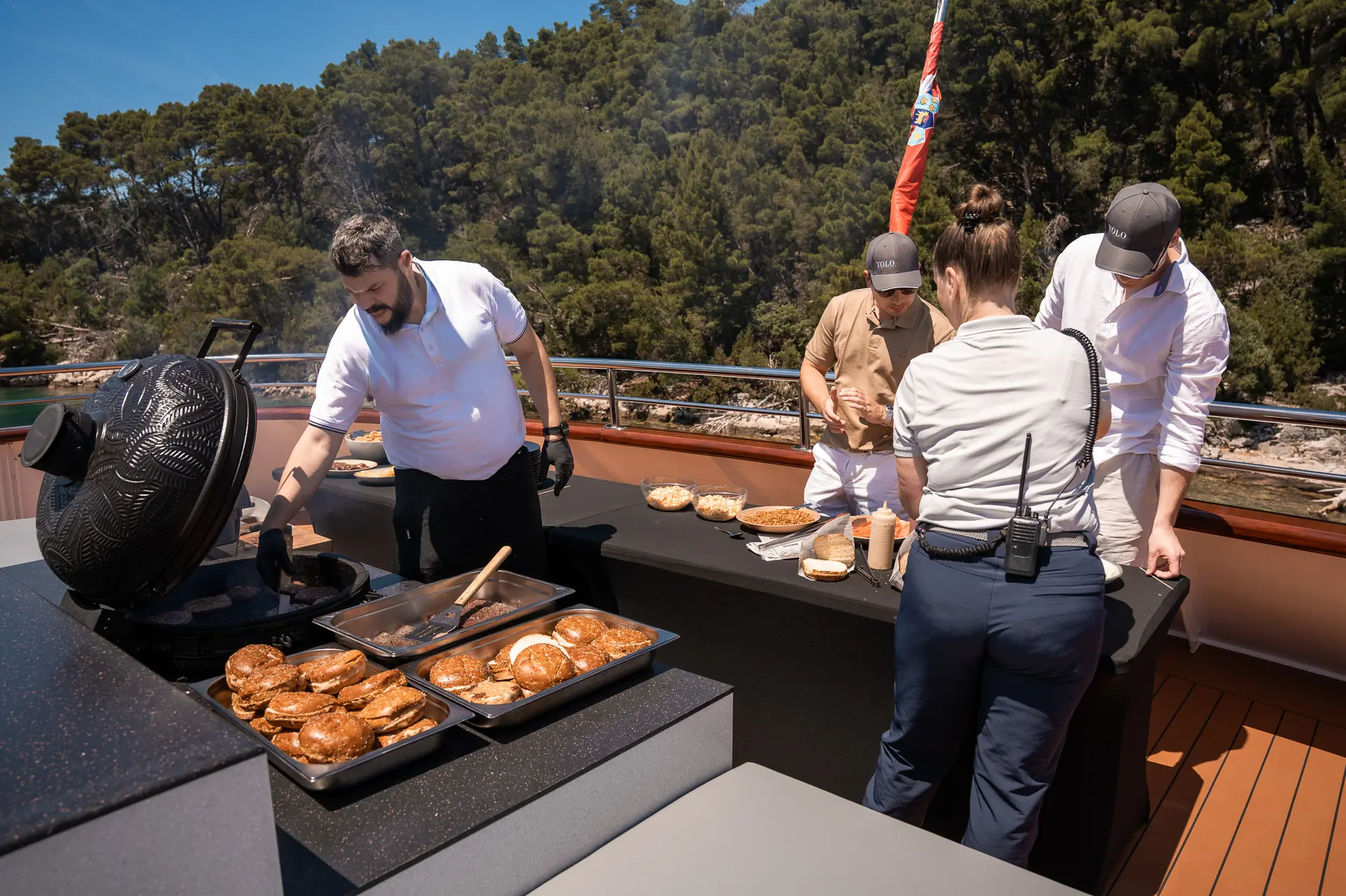 Four people are preparing food outdoors on a yacht, with burgers and buns cooking on a grill and additional food on tables. Trees and greenery are visible in the background.