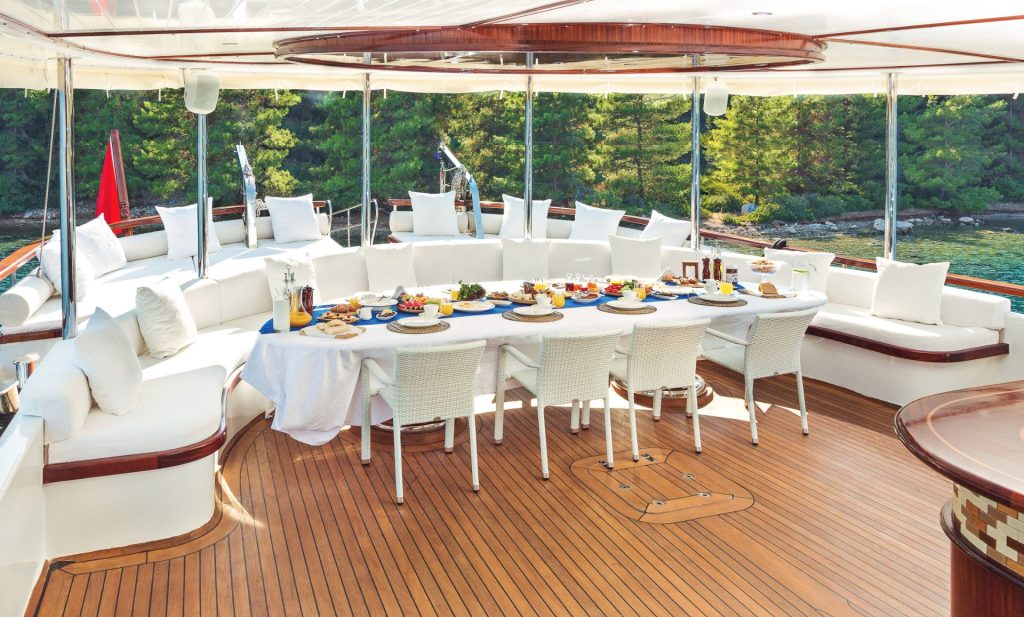A large outdoor dining table set for a meal, surrounded by white chairs and a curved white cushioned sofa on a yacht deck, with trees and water visible in the background.