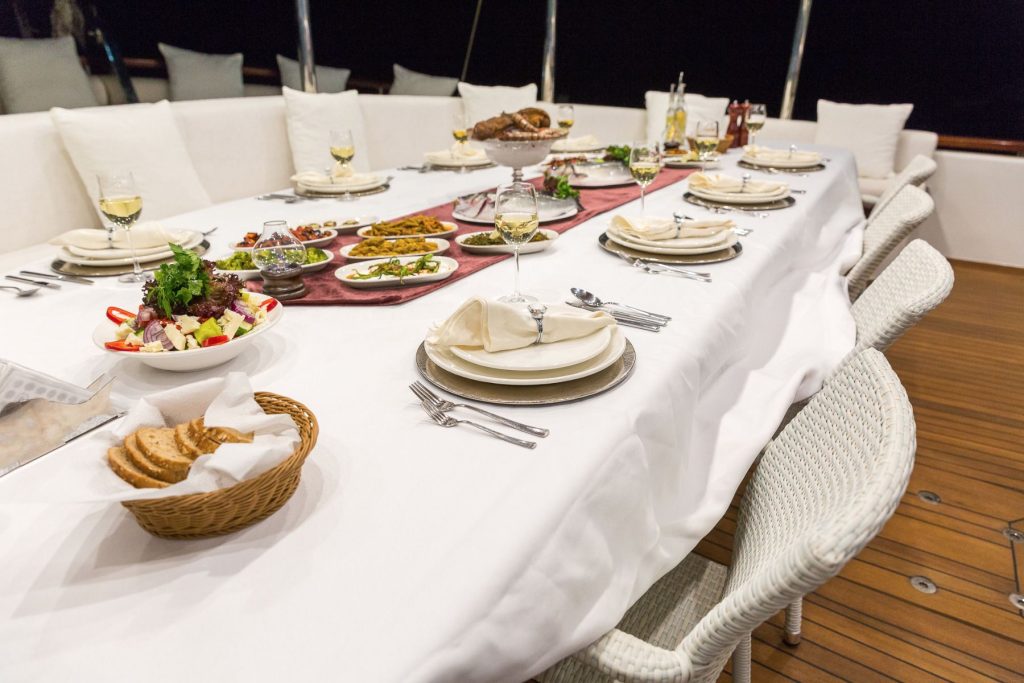 A long, white-clothed dining table set for several guests, with plates, cutlery, wine glasses, salads, bread, and various dishes, on a wooden deck with cushioned white chairs and a dark background.