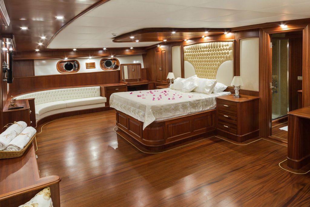 Luxurious yacht bedroom with polished wood floors and walls, a large bed decorated with rose petals, a cream tufted headboard, white sofa, bedside tables with lamps, and round porthole windows.