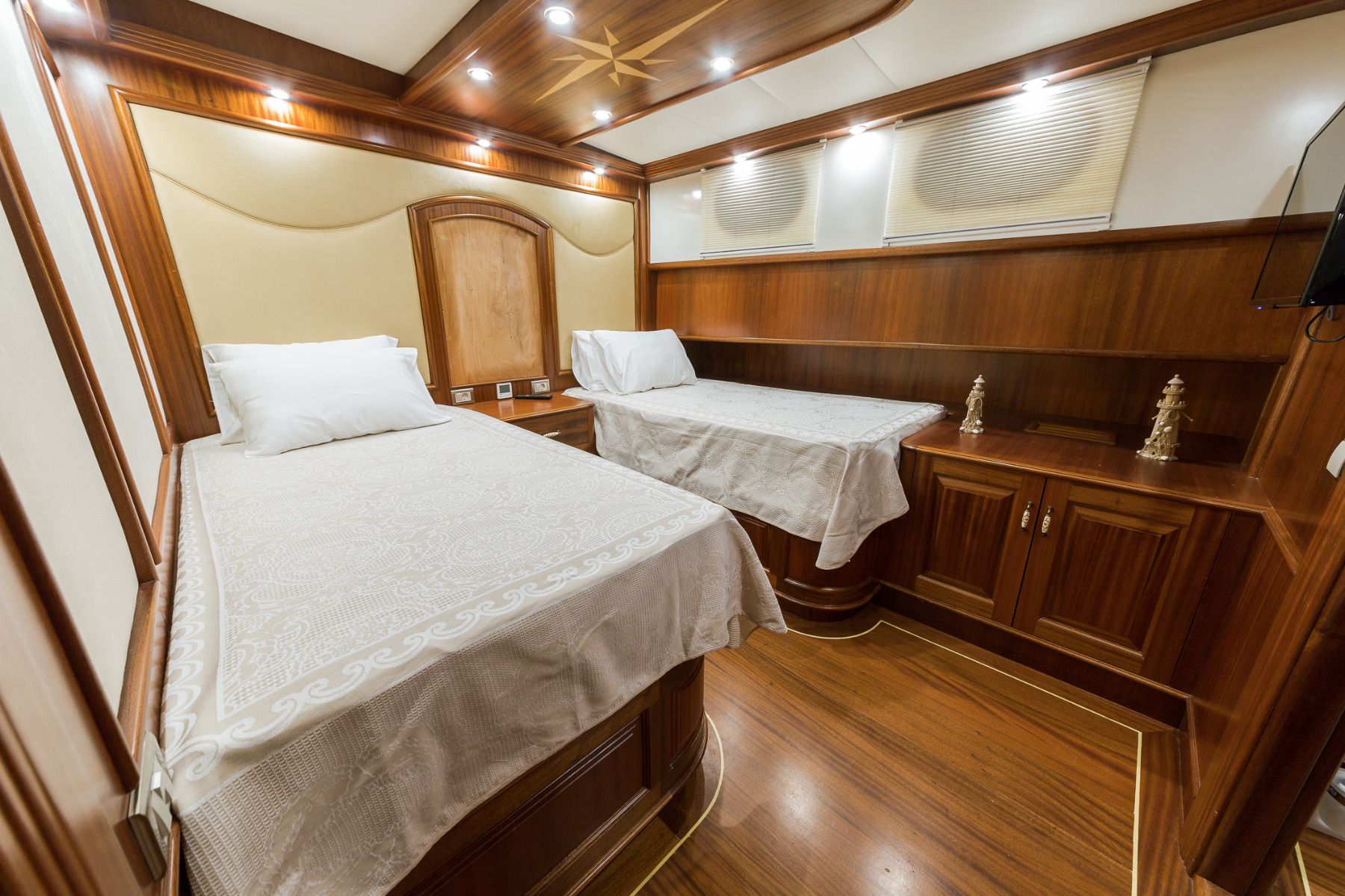 A yacht cabin with polished wood walls and floors, featuring two single beds with white linens, built-in cabinets, decorative chess pieces, and soft overhead lighting.