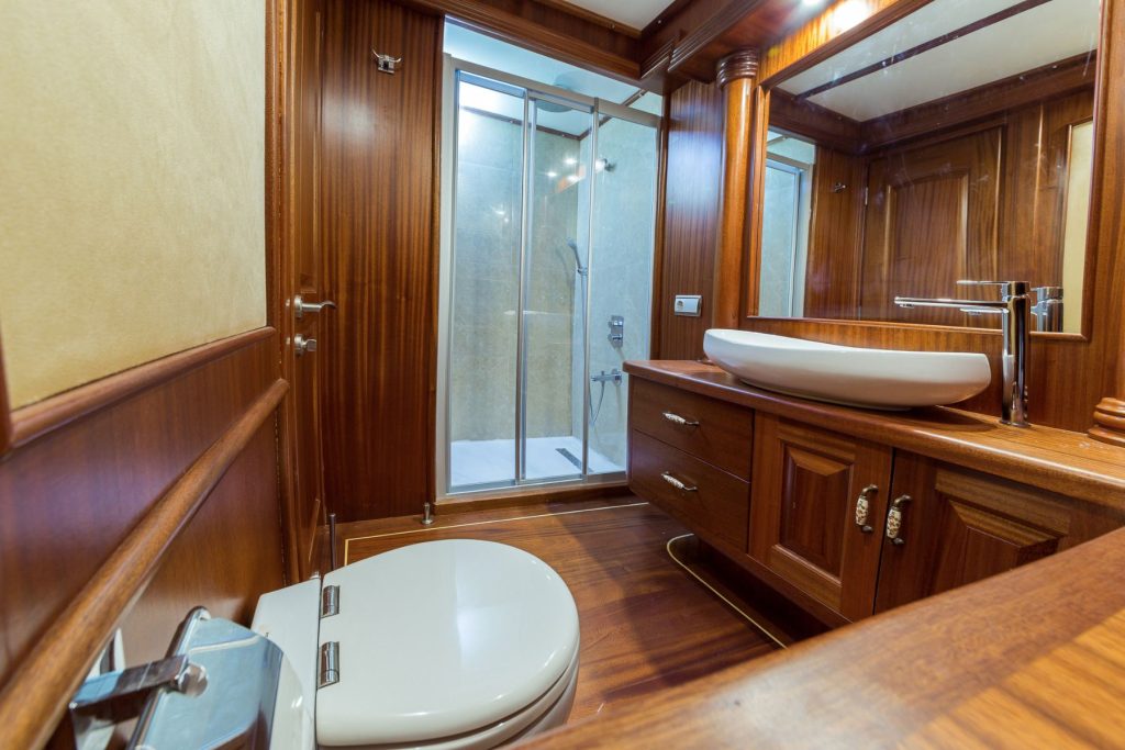 A luxurious bathroom with wooden walls and floor, featuring a modern toilet, a large sink with a cabinet, a wide mirror, and a glass-enclosed shower in the background.