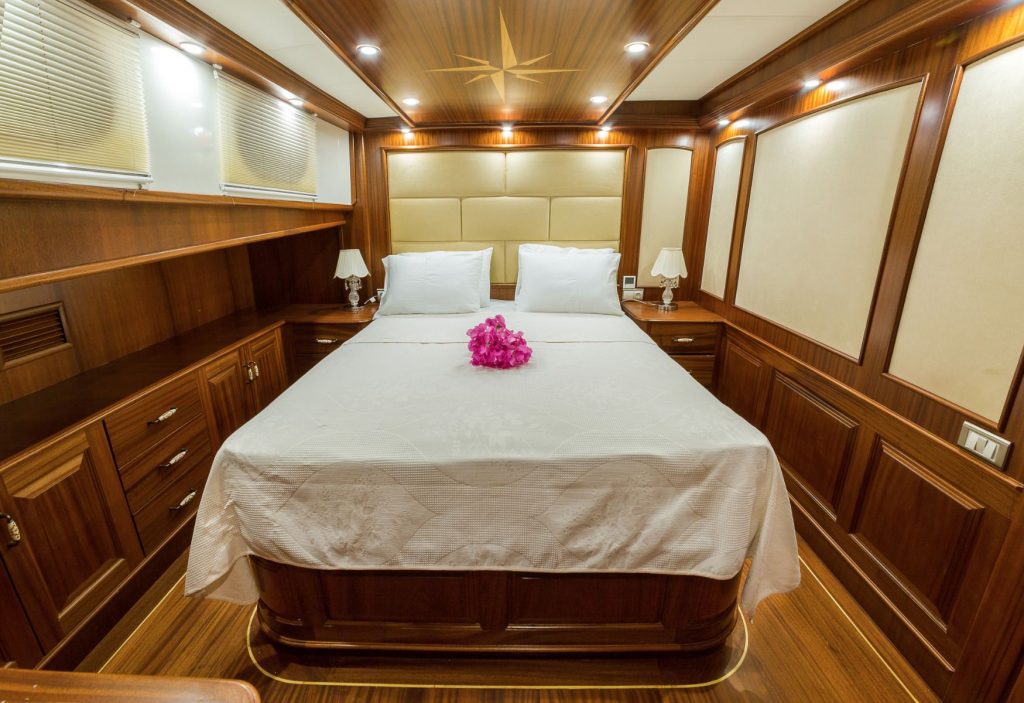 Luxurious yacht cabin with wood-paneled walls, a double bed with white linens and pillows, two bedside lamps, and a bouquet of pink flowers on the bed. Warm lighting creates a cozy atmosphere.