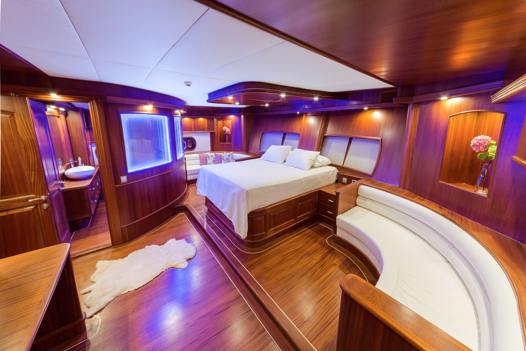 Luxurious yacht bedroom with polished wood walls, a large bed, curved white sofa, soft lighting, fur rug, and flowers in a vase, creating an elegant and inviting atmosphere.
