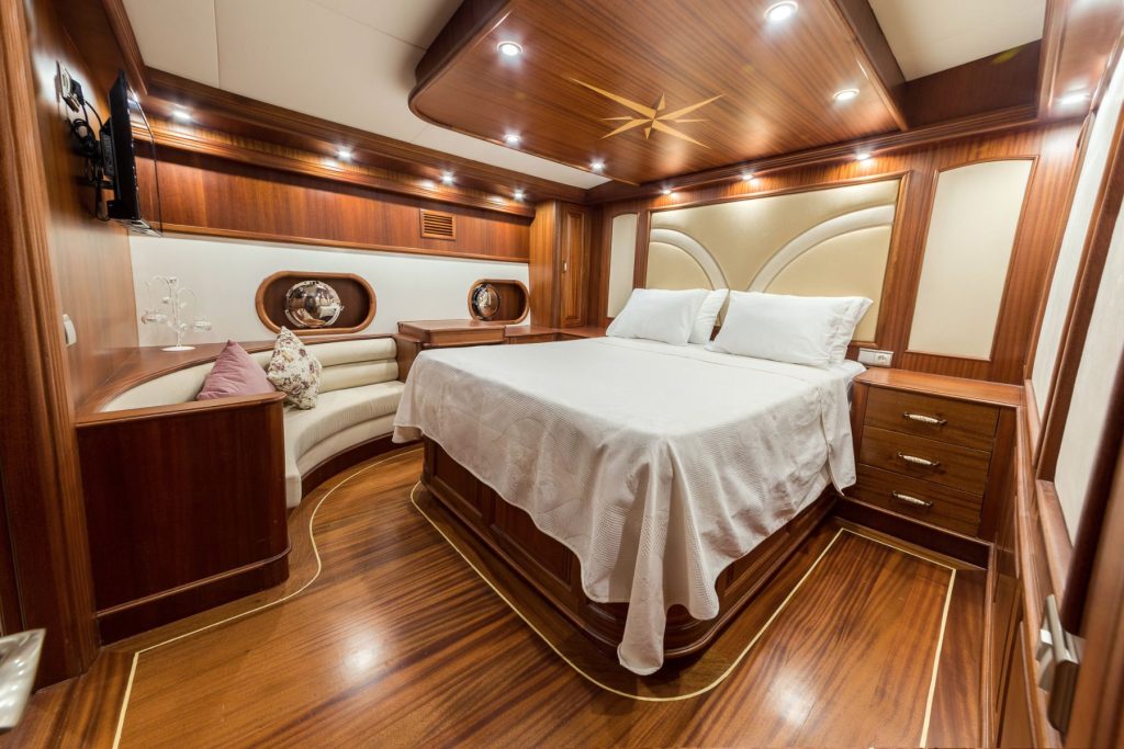Luxurious yacht bedroom with rich wood paneling, a large bed with white linens, built-in shelves, two bedside tables, soft lighting, and a cozy curved seating area with cushions.