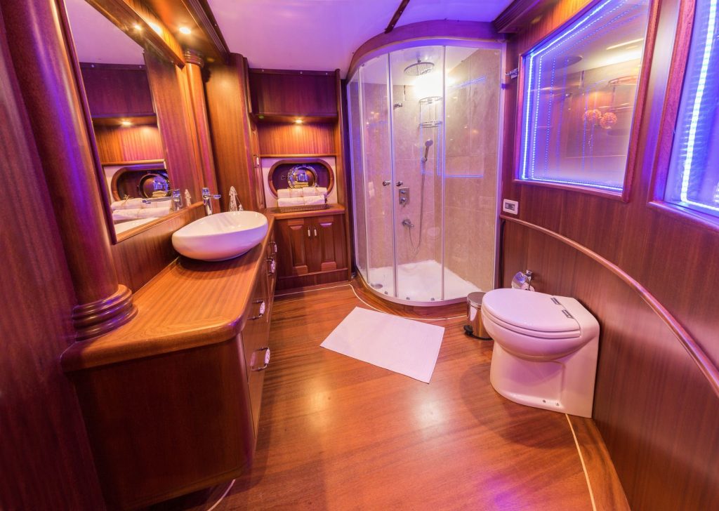 Luxurious bathroom with wood-paneled walls and floors, a modern glass shower, white sink, toilet, large mirror with blue LED lighting, and warm ambient lighting throughout the space.