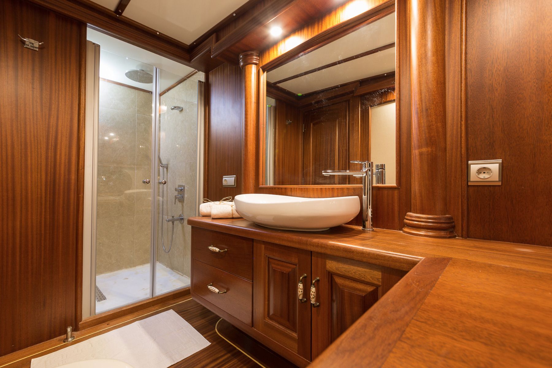 Luxurious bathroom with rich wood paneling, a modern white vessel sink on a wooden counter, large mirror, and a glass-enclosed shower with beige tiles. The space is well-lit and elegant.