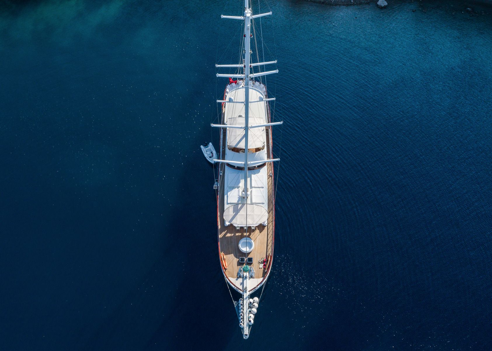 A large sailboat seen from above, floating on calm, deep blue water near a shoreline. The boat has multiple masts and a wooden deck, with a small tender boat tied alongside.