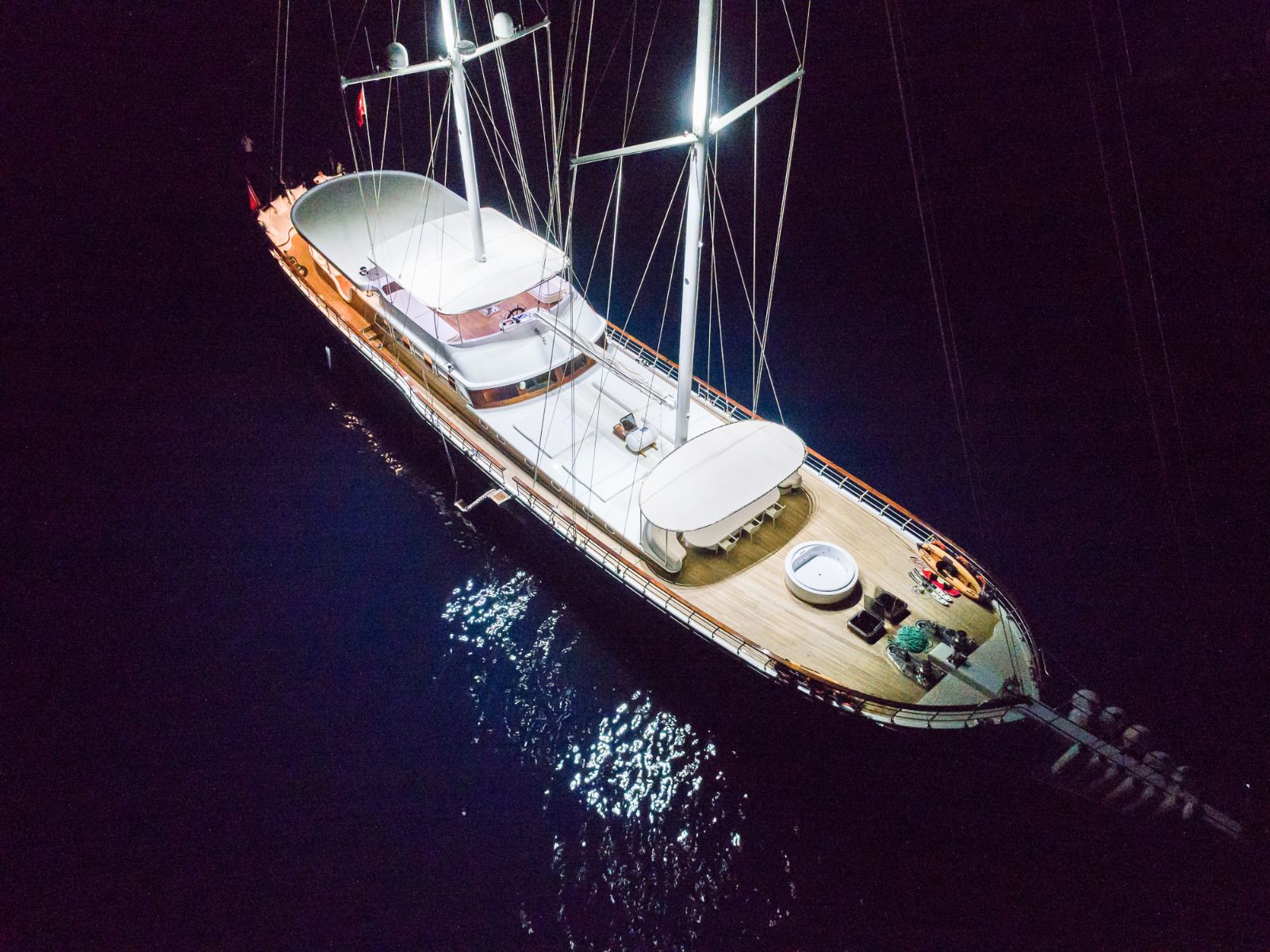 A large, elegant sailboat is illuminated at night, floating on dark, calm water. The deck is well-lit, revealing seating areas, a hot tub, and visible masts and rigging, with light reflecting on the water’s surface.