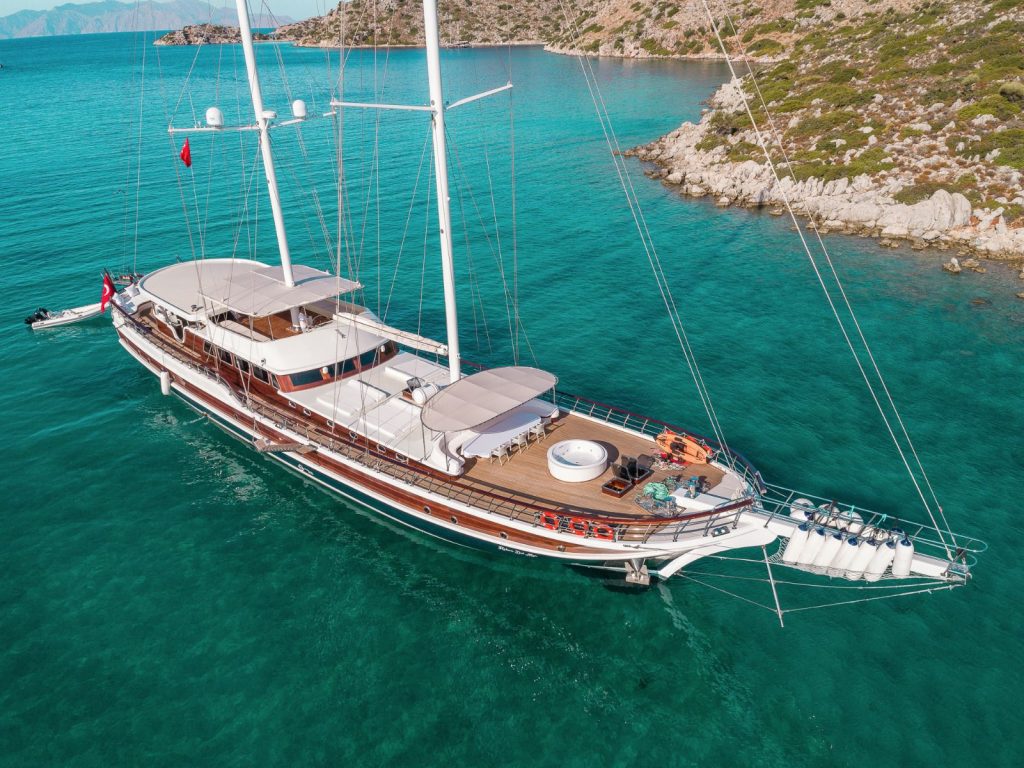 A large, luxurious sailing yacht with wooden decking and white canopies is anchored in clear turquoise water near a rocky, green coastline. Sun loungers, a jacuzzi, and outdoor seating are visible on the deck.