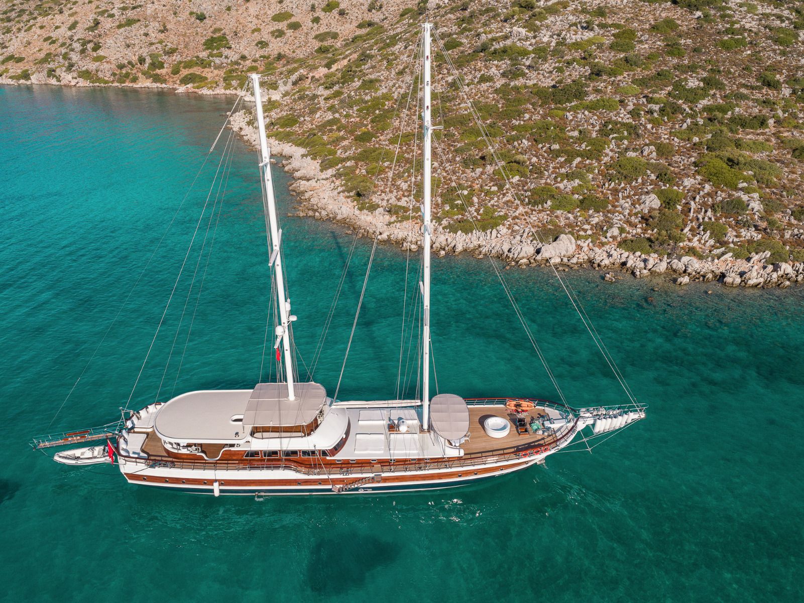 A large sailboat with two masts floats on clear turquoise water near a rocky, shrub-covered coastline under a sunny sky.