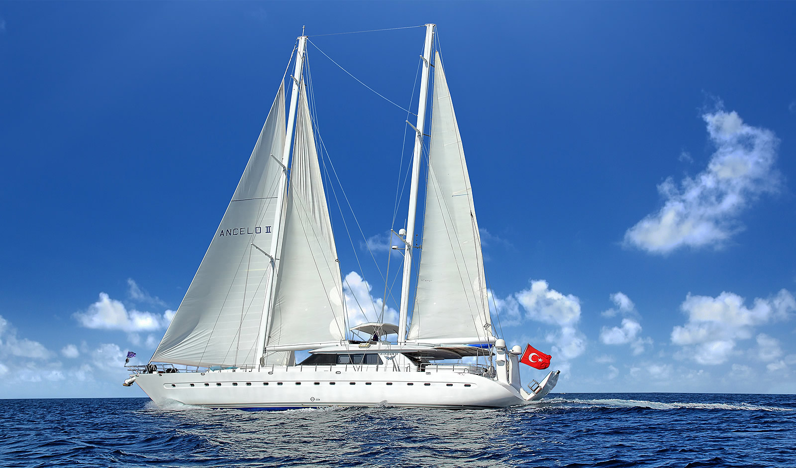 ANGELO II A large white sailing yacht with two tall masts moves across calm blue water under a clear sky. The yacht displays a red Turkish flag at the stern. Fluffy white clouds are scattered across the background.