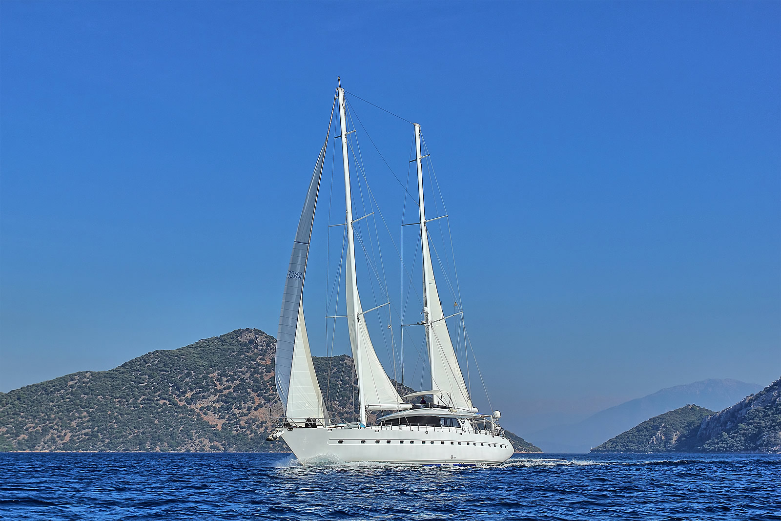 ANGELO II A large white sailboat with three masts sails on calm blue water, with hilly, green coastline and clear blue sky in the background.