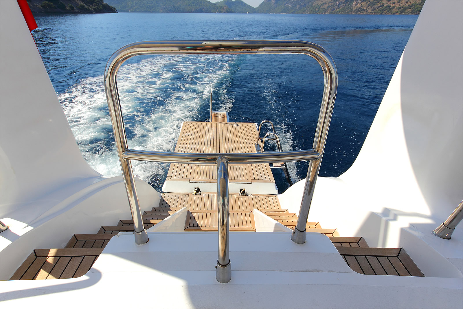 ANGELO II View from the deck of a yacht looking out over the water, with wooden steps and a metal railing leading down to a swim platform at the back of the boat; the sea and distant hills are visible.