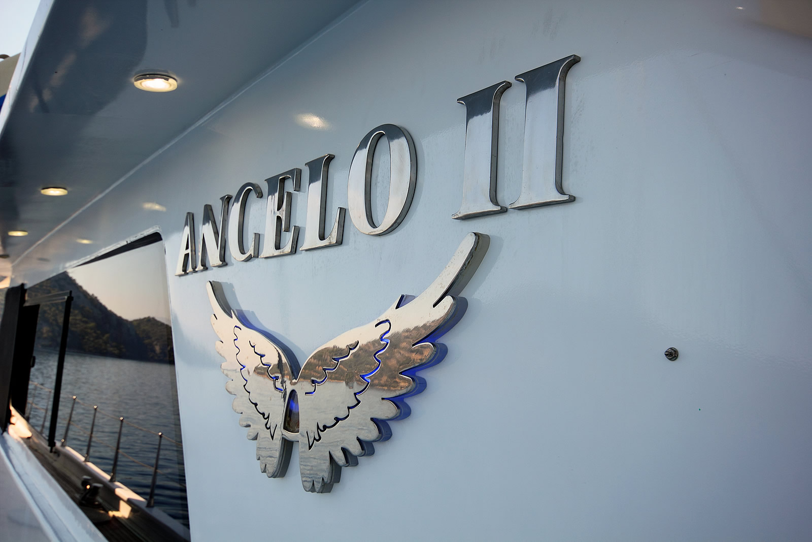 ANGELO II The side of a yacht with the name Angelo II and a metallic angel wings emblem; reflections of water and distant mountains are visible in the background.