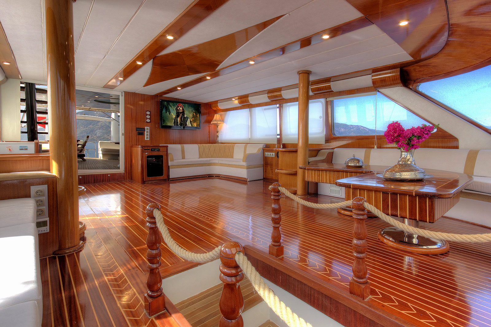 ANGELO II Luxurious yacht interior with polished wood floors and ceilings, cream-colored seating, a large coffee table with a vase of pink flowers, and a flat-screen TV; large windows offer scenic water views.