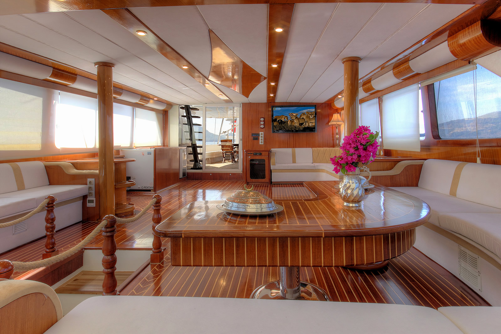 ANGELO II Spacious yacht interior with polished wood floors and pillars, cream-colored sofas, a round table with a vase of pink flowers, large windows, and a flat-screen TV on the wall. Sunlight streams in through the windows.