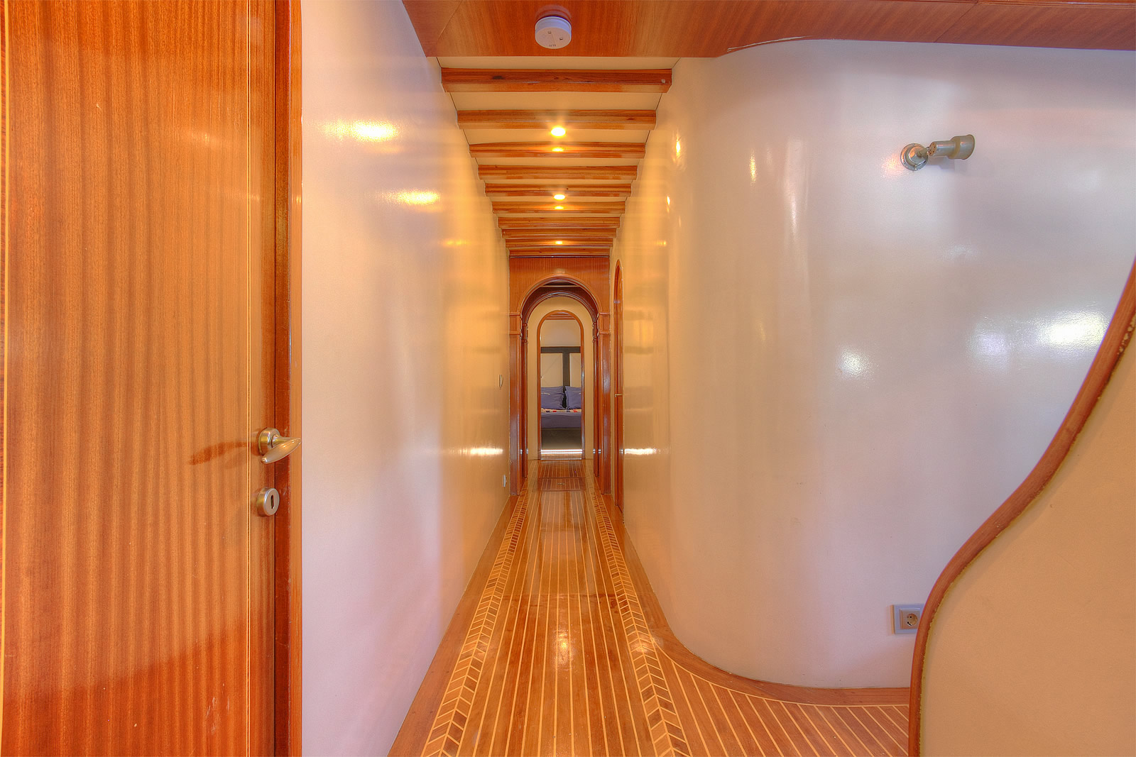 ANGELO II A narrow, well-lit hallway with wooden floors and walls, featuring curved white surfaces and multiple doorways leading toward a room at the end. The ceiling has exposed wood beams and recessed lighting.