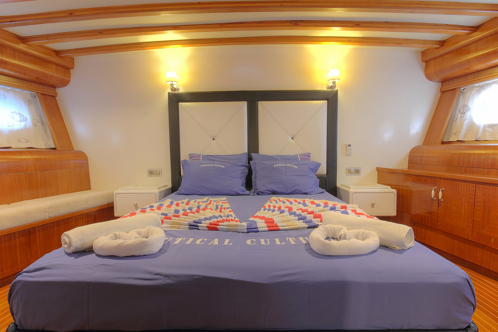 ANGELO II A cozy, well-lit yacht cabin with a double bed covered in blue bedding, two rolled towels at the foot, wood-paneled walls and ceiling, small windows with curtains, and bedside wall lamps.
