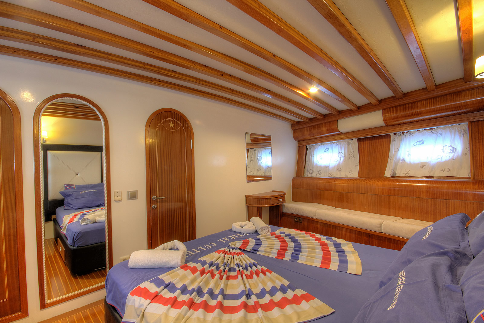 ANGELO II A cozy yacht cabin with wooden walls and ceiling beams, a double bed with blue linens, towels folded in fan shapes, a mirror, a door, a small desk, and a cushioned bench under curtained windows.