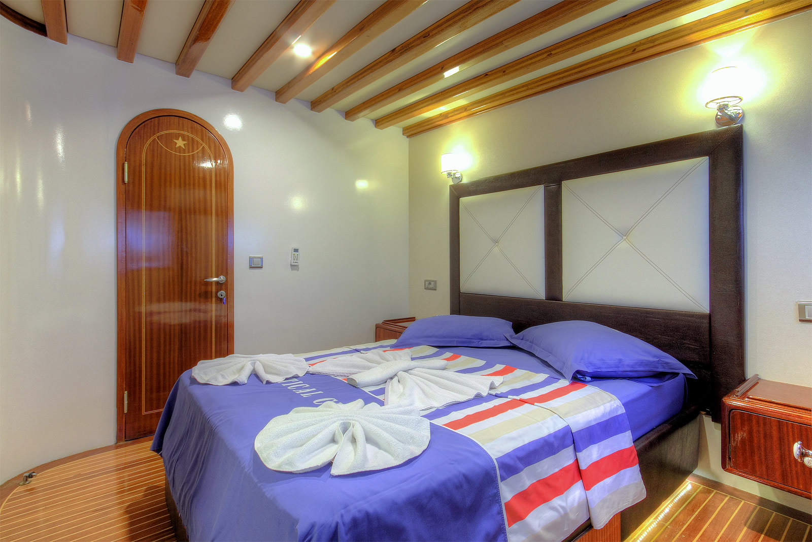 ANGELO II A cozy bedroom with a wooden door, double bed with blue and striped bedding, two towel swans and heart shapes on the bed, wooden ceiling beams, and two wall-mounted lights above the headboard.