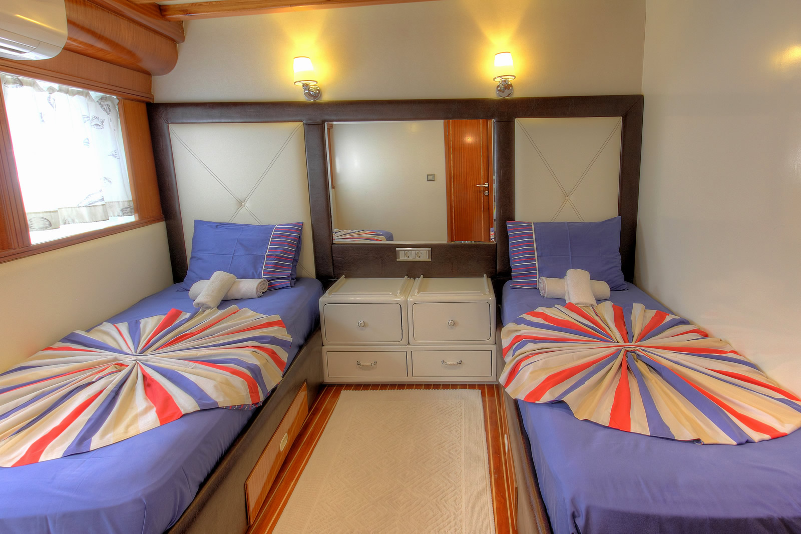 ANGELO II A cozy bedroom with two single beds covered in blue sheets and colorful, fan-shaped duvets. Each bed has a rolled towel, and a small cabinet with drawers sits between them beneath a large mirror.